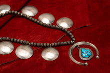 Load image into Gallery viewer, A Navajo squash blossom necklace with a large turquoise stone at the center, featuring large round silver metalwork, displayed against red leather background.