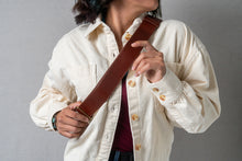 Load image into Gallery viewer, Person wearing a creme colored button shirts holds the brown leather belt strap of a small backpack