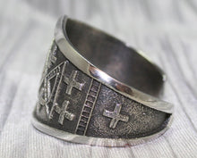 Load image into Gallery viewer, handcrafted silver cuff bracelet featuring intricate geometric and cross patterns etched into the metal. The design is bold and symmetrical, with a textured background that enhances the detailed craftsmanship. The bracelet rests on a neutral surface, emphasizing its artistic and traditional appeal.
