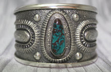 Load image into Gallery viewer, A bold sand-cast cuff featuring intricate textures and patterns, with a vibrant oval Pilot Mountain turquoise stone at the center. The turquoise showcases deep blue tones with a coppery matrix, framed by an ornate border that adds depth and elegance. The design reflects a balance of rugged craftsmanship and refined artistry.