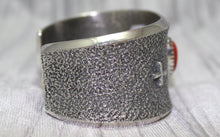 Load image into Gallery viewer, A wide silver cuff bracelet with a textured surface, small silver cross on side. The cuff rests on a soft gray background, highlighting its intricate craftsmanship.