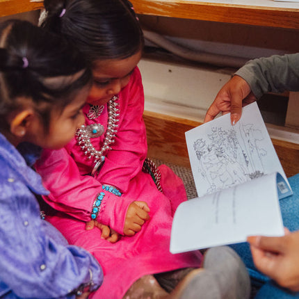 Two little Navajo girls wearing traditional clothing are looking at a children's book that a older Navajo girl is reading to them