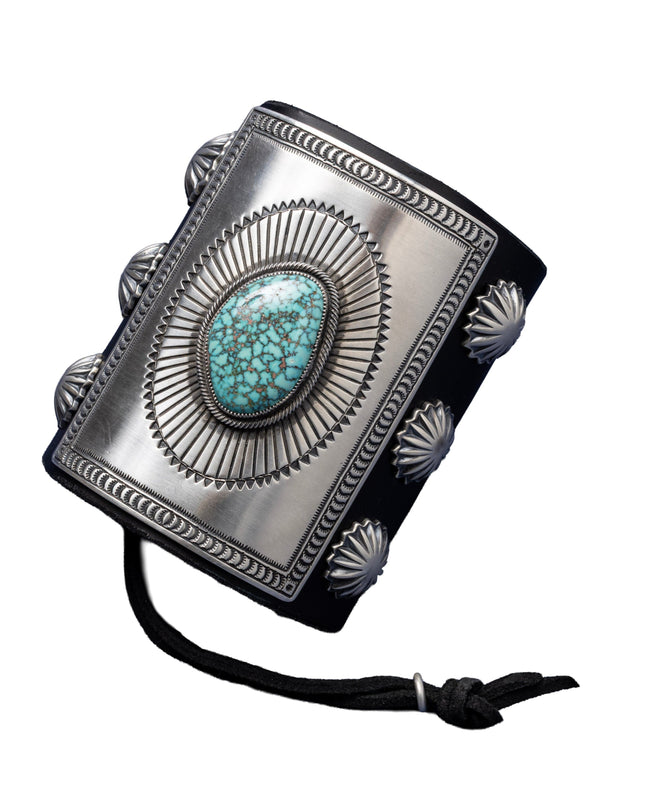 A bold leather bowguard adorned with a rectangular sterling silver faceplate, finely stamped in a sunburst motif framing a sky-blue Kingman turquoise stone with dark matrix. Embellished with fan-pattern silver conchos on both sides and tied with a black leather cord.