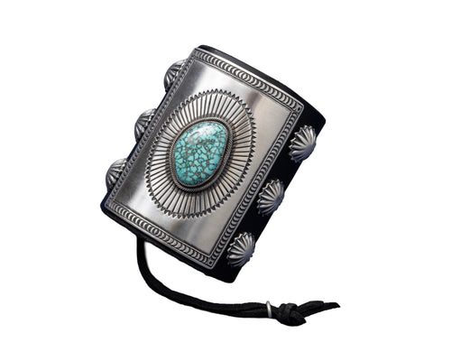 A bold leather bowguard adorned with a rectangular sterling silver faceplate, finely stamped in a sunburst motif framing a sky-blue Kingman turquoise stone with dark matrix. Embellished with fan-pattern silver conchos on both sides and tied with a black leather cord.