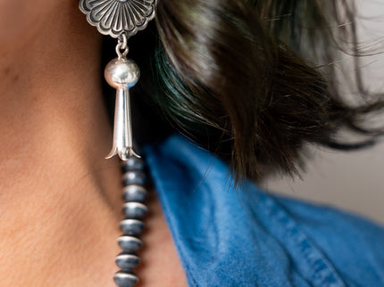 Close-up of a circular silver drop earring with a silver tassel hanging from the ear of a Navajo woman wearing a blue shirt.