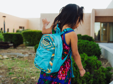Little Navajo girl wearing a colorful dress and blue Pendleton backpack in front of a college building