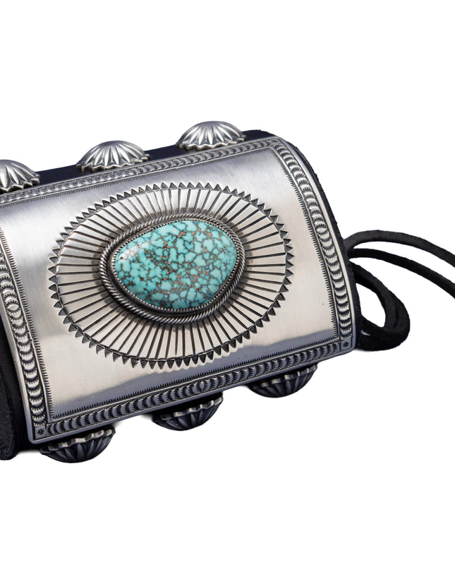 A bold leather bowguard adorned with a rectangular sterling silver faceplate, finely stamped in a sunburst motif framing a sky-blue Kingman turquoise stone with dark matrix. Embellished with fan-pattern silver conchos on both sides and tied with a black leather cord.
