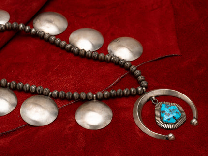 A Navajo squash blossom necklace with a large turquoise stone at the center, featuring large round silver metalwork, displayed against red leather background.