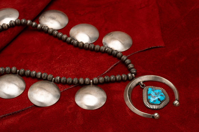 A Navajo squash blossom necklace with a large turquoise stone at the center, featuring large round silver metalwork, displayed against red leather background.