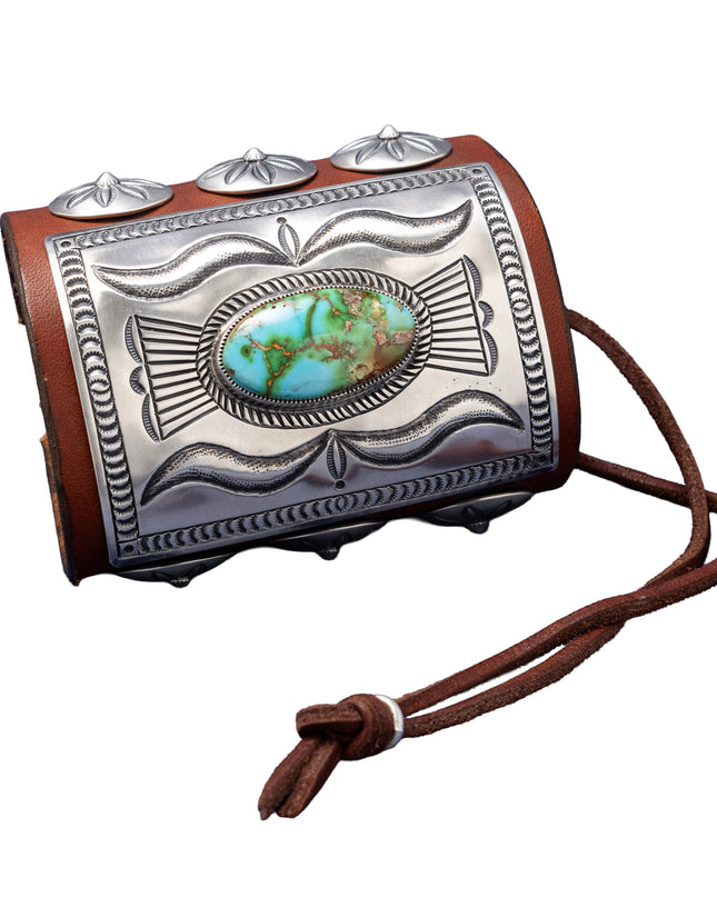 A richly detailed leather bowguard with a rectangular sterling silver plate showcasing flowing stamped patterns and a striking green-blue Sonoran Gold turquoise stone at the center. Decorated with dome-shaped silver conchos and secured with a dark brown leather tie.