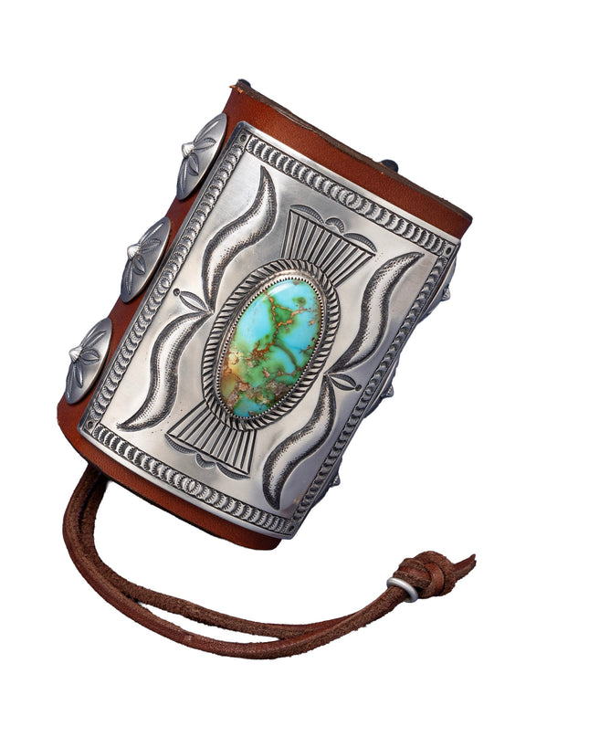A richly detailed leather bowguard with a rectangular sterling silver plate showcasing flowing stamped patterns and a striking green-blue Sonoran Gold turquoise stone at the center. Decorated with dome-shaped silver conchos and secured with a dark brown leather tie.