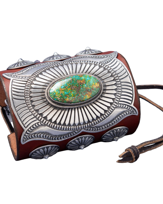 A handcrafted leather bowguard featuring an elaborate stamped sterling silver overlay with a radiant sunburst design, centered around a vibrant Sonoran Gold turquoise cabochon. Accented with scalloped conchos on the sides and a brown leather strap for fastening.