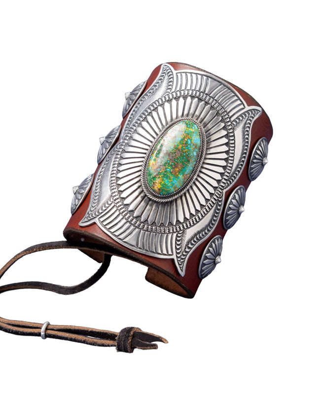 A handcrafted leather bowguard featuring an elaborate stamped sterling silver overlay with a radiant sunburst design, centered around a vibrant Sonoran Gold turquoise cabochon. Accented with scalloped conchos on the sides and a brown leather strap for fastening.