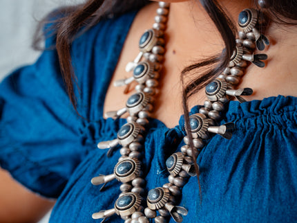 Navajo woman wearing a blue dress and a detailed Navajo squash blossom necklace with blue gem stones.
