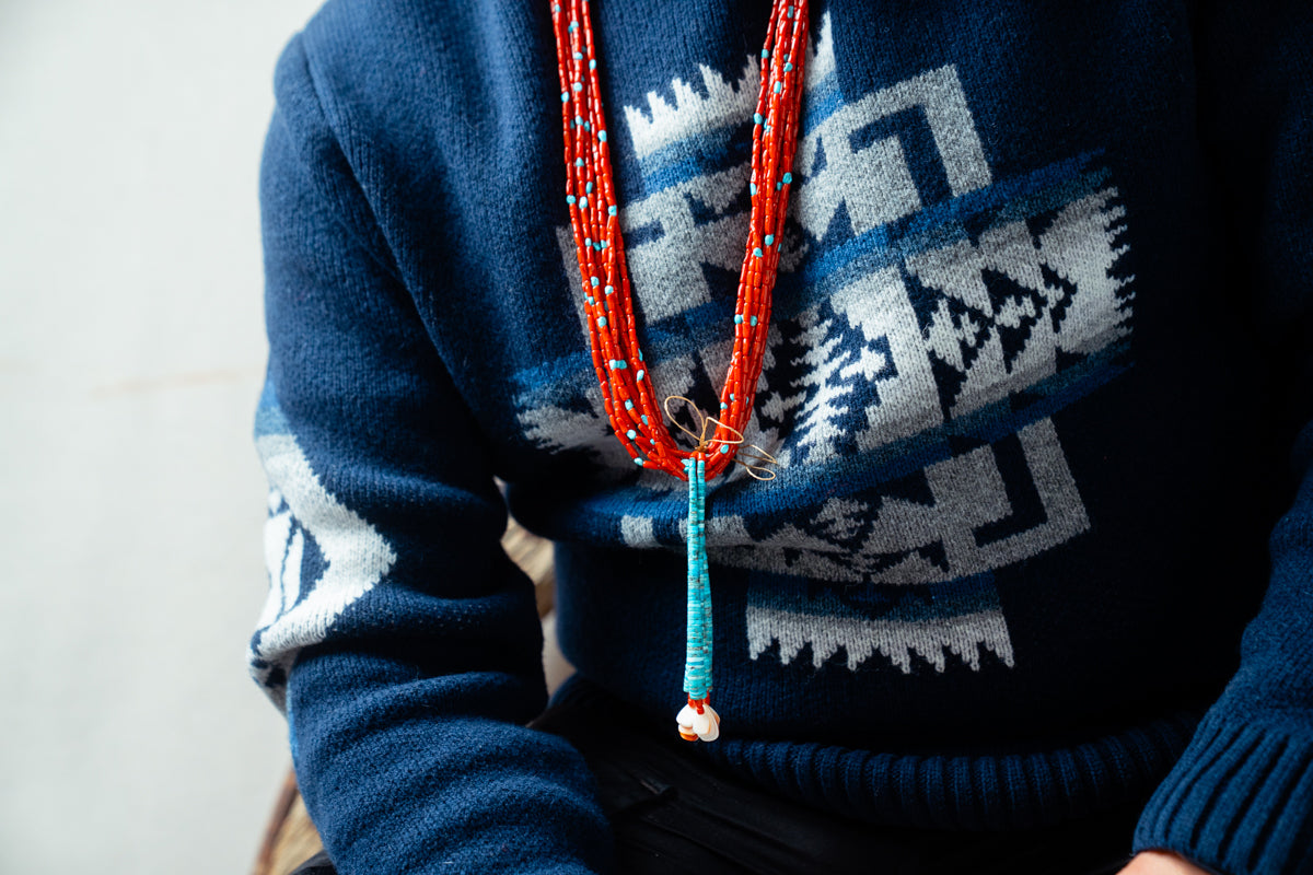Person wearing a blue Pendleton sweater with a coral jacloo necklace.