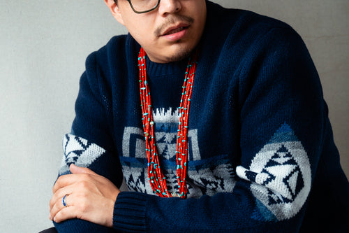 Man wearing a dark blue Pendleton sweater with geometric patterns and a red coral necklace against a gray background