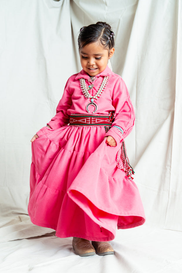 Navajo little girl wearing a traditional pink dress with a red sash belt and small silver squash blossom on a tan background