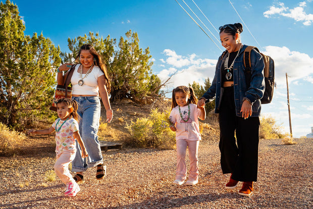 Two Navajo women walking on a dirt road with two Navajo children wearing modern clothing and Native American jewelry.