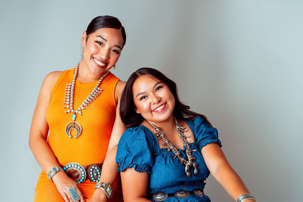 Two smiling Navajo women leaning on each other wearing squash blossoms and other native American jewelry against a tan background