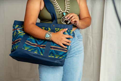 Navajo women in modern clothes has a Pendleton on their right shoulder and holding a Navajo squash blossom with their left hand