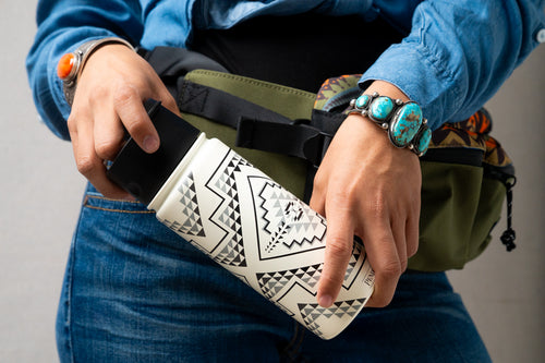 Close up of a person holding a white Pendleton patterned mug at waist level with a green fanny pack behind the mug