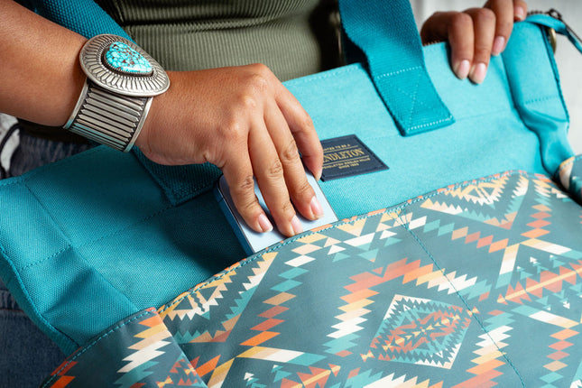 Close up of a females's right hand placing a blue smart phone into the outside pocket  of a Pendleton tote bag. Model is wearing a large turquoise bracelet on her wrist.