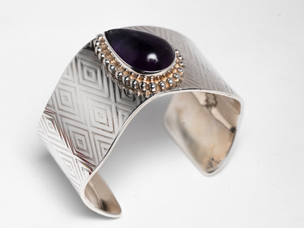A silver bracelet with diamond-shaped stamped patterns and a large deep purple sugalite stone, featuring a chain of smaller beads.
