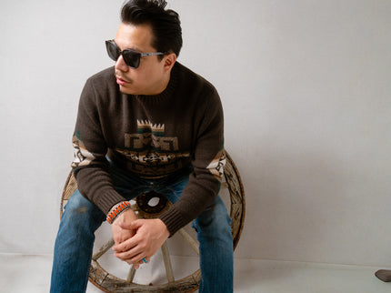 Navajo male sitting on a wooden wheel wearing Pendleton sunglasses and brown knitted sweater with blue jeans