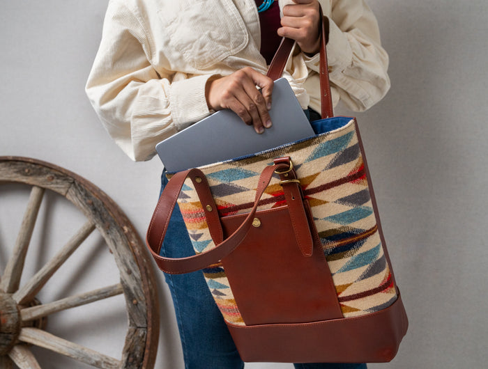 Women puling out a silver MacBook Pro with one hand while holding a colorful and brown leather Pendleton bag with a rustic wheel in the background