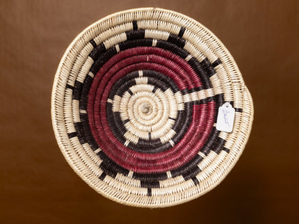 A vintage Navajo basket with a starburst pattern in natural, red, and black tones, shown against a warm brown background.
