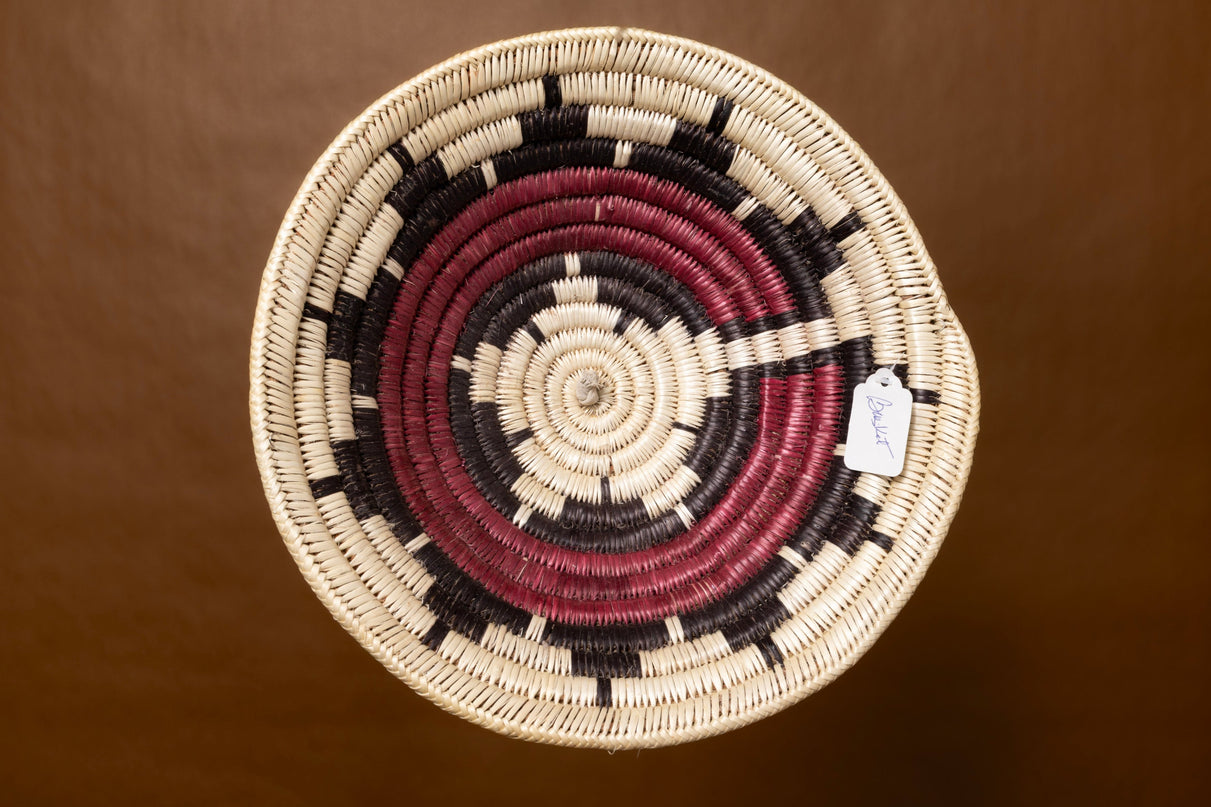 A vintage Navajo basket with a starburst pattern in natural, red, and black tones, shown against a warm brown background.