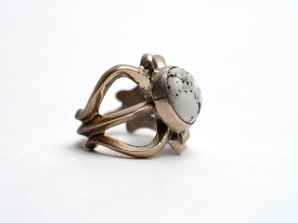 Profile view of silver cast cut-out ring showcases a single, centrally set round white buffalo stone on a white background. Ring appears gold because of the natural patina