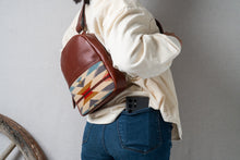 Load image into Gallery viewer, Navajo woman wearing a brown leather backpack with a geometric woven pattern, with a phone in back pants pocket, against a creme background.

