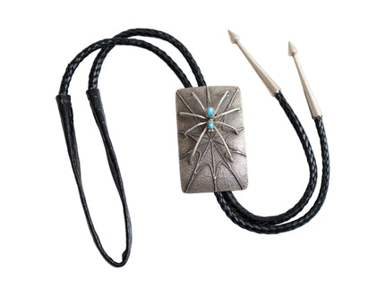 Full view of a bolo tie that has the design of silver spider with two turquoise stones as the body raised on a rectangular silver Bolo tie with spider web etching with a black leather cord and two silver tips