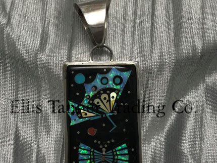 A rectangular pendant with a silver frame and loop for a chain. The pendant features an intricate design with geometric patterns in vibrant turquoise, black, and yellow, resembling a butterfly and spreading it wings. The background has shimmering details, creating a striking contrast.