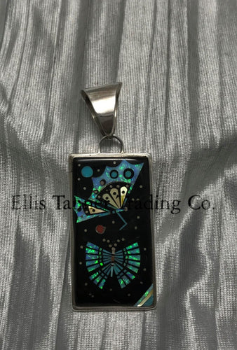 A rectangular pendant with a silver frame and loop for a chain. The pendant features an intricate design with geometric patterns in vibrant turquoise, black, and yellow, resembling a butterfly and spreading it wings. The background has shimmering details, creating a striking contrast.