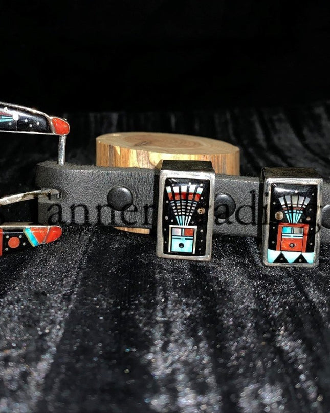 A handcrafted Ranger belt buckle set featuring intricate inlay work. The set includes a buckle, two keepers, and a belt tip. The design incorporates vibrant geometric patterns with black, turquoise, coral, and white inlays, inspired by Southwestern artistry. The bold and colorful patterns are framed by polished silver, enhancing the intricate details. The set is displayed on a black leather belt, resting on a textured dark background.