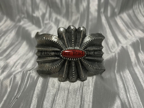 A silver-colored cuff bracelet with a central coral stone, on a textured silver background.