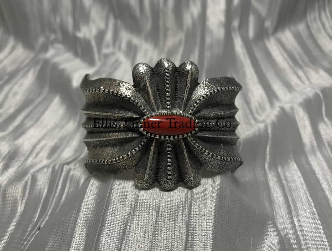 A silver-colored cuff bracelet with a central coral stone, on a textured silver background.
