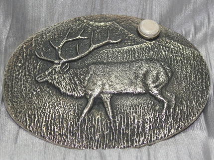A sand-cast buckle featuring an intricately etched elk design set against a textured background. Added is a small, antler-shaped stone, adding a distinctive and natural touch. The highlighting its rustic and artisanal craftsmanship.