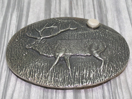 A sand-cast buckle featuring an intricately etched elk design set against a textured background. Added is a small, antler-shaped stone, adding a distinctive and natural touch. The highlighting its rustic and artisanal craftsmanship.