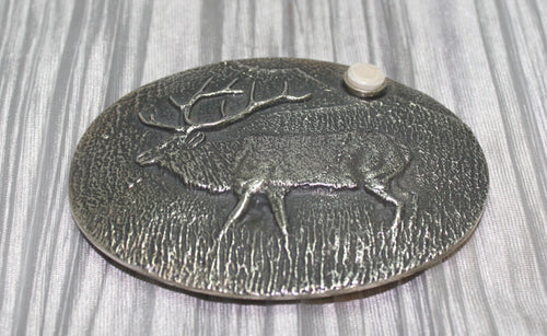 A sand-cast buckle featuring an intricately etched elk design set against a textured background. Added is a small, antler-shaped stone, adding a distinctive and natural touch. The highlighting its rustic and artisanal craftsmanship.