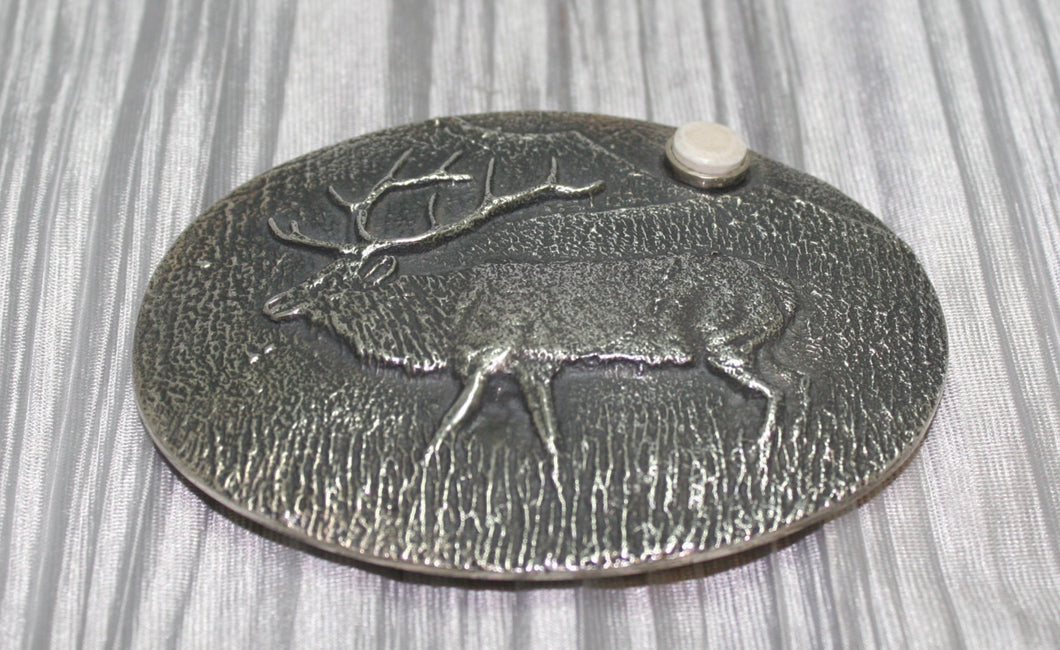 A sand-cast buckle featuring an intricately etched elk design set against a textured background. Added is a small, antler-shaped stone, adding a distinctive and natural touch. The highlighting its rustic and artisanal craftsmanship.