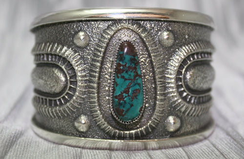 A bold sand-cast cuff featuring intricate textures and patterns, with a vibrant oval Pilot Mountain turquoise stone at the center. The turquoise showcases deep blue tones with a coppery matrix, framed by an ornate border that adds depth and elegance. The design reflects a balance of rugged craftsmanship and refined artistry.