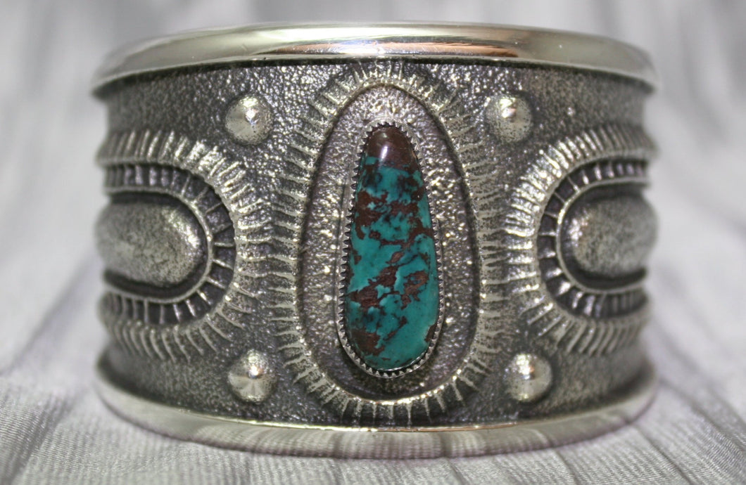 A bold sand-cast cuff featuring intricate textures and patterns, with a vibrant oval Pilot Mountain turquoise stone at the center. The turquoise showcases deep blue tones with a coppery matrix, framed by an ornate border that adds depth and elegance. The design reflects a balance of rugged craftsmanship and refined artistry.