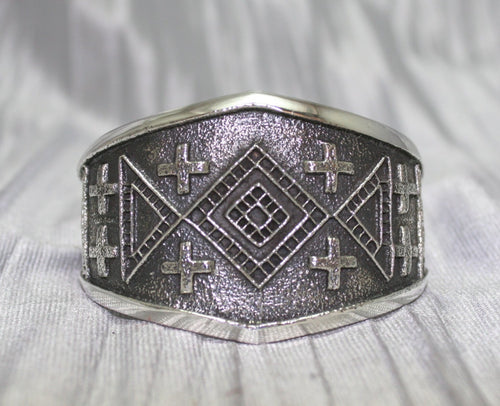 handcrafted silver cuff bracelet featuring intricate geometric and cross patterns etched into the metal. The design is bold and symmetrical, with a textured background that enhances the detailed craftsmanship. The bracelet rests on a neutral surface, emphasizing its artistic and traditional appeal.