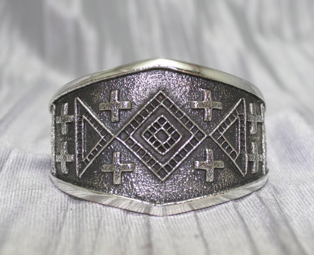 handcrafted silver cuff bracelet featuring intricate geometric and cross patterns etched into the metal. The design is bold and symmetrical, with a textured background that enhances the detailed craftsmanship. The bracelet rests on a neutral surface, emphasizing its artistic and traditional appeal.