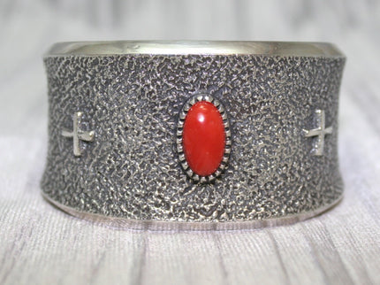 A wide silver cuff bracelet with a textured surface, featuring a bright oval red stone at the center and two small silver crosses on each side. The cuff rests on a soft gray background, highlighting its intricate craftsmanship.