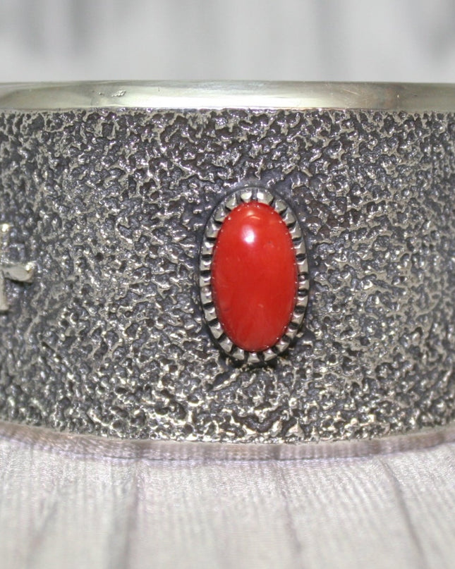 A wide silver cuff bracelet with a textured surface, featuring a bright oval red stone at the center and two small silver crosses on each side. The cuff rests on a soft gray background, highlighting its intricate craftsmanship.