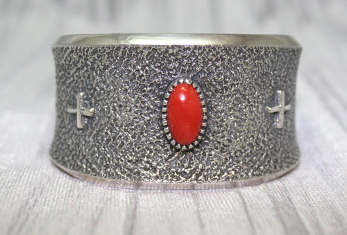 A wide silver cuff bracelet with a textured surface, featuring a bright oval red stone at the center and two small silver crosses on each side. The cuff rests on a soft gray background, highlighting its intricate craftsmanship.
